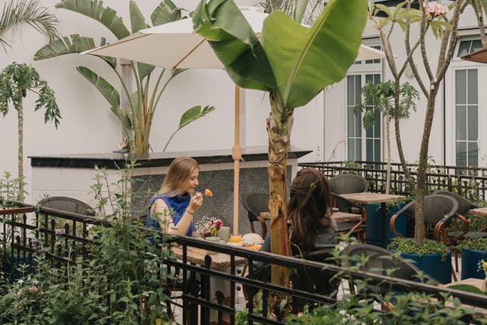 Two guests have breakfast on the rooftop garden terrace, among banana plants and under a parasol