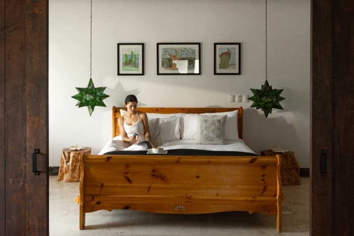 A room with a carved wooden bed, green star-shaped lamps and three cactus watercolours above the headboard; someone eats breakfast sitting on the bed