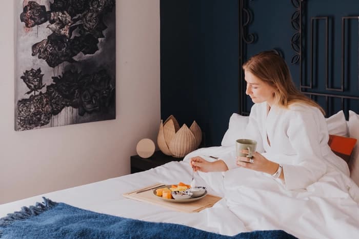 Someone in a white robe having breakfast in bed from a tray, beside a blue wall and a painting of dark flowers
