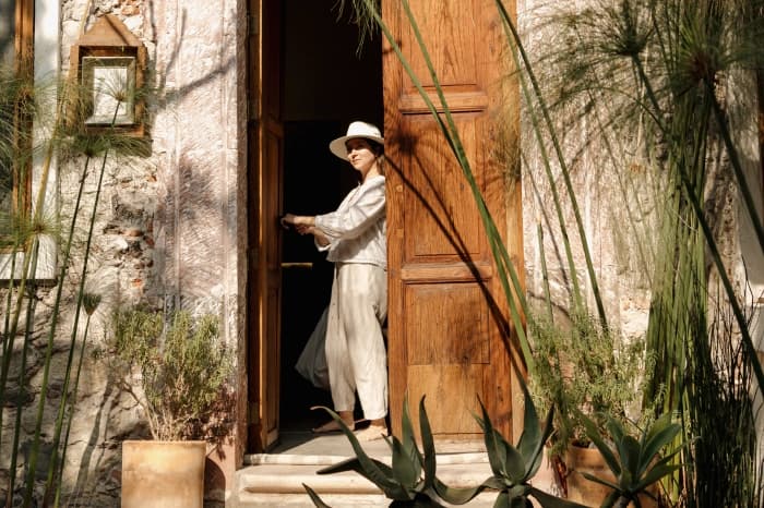 Someone in light linen and a brimmed hat in the wooden doorway, among papyrus and agaves