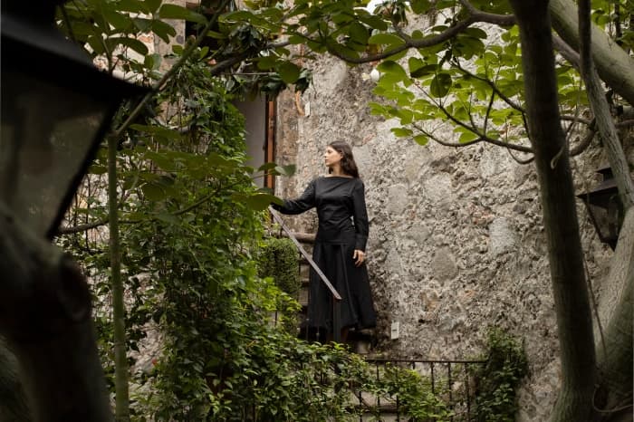 Someone in a long dress on a stair of the house, between the stone walls and the branches of the courtyard