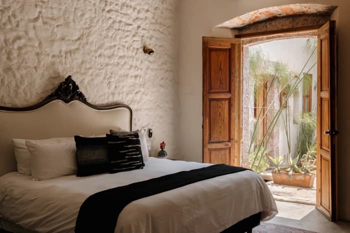 A room with whitewashed stone walls, a carved headboard, dark cushions and the wooden door open to the courtyard