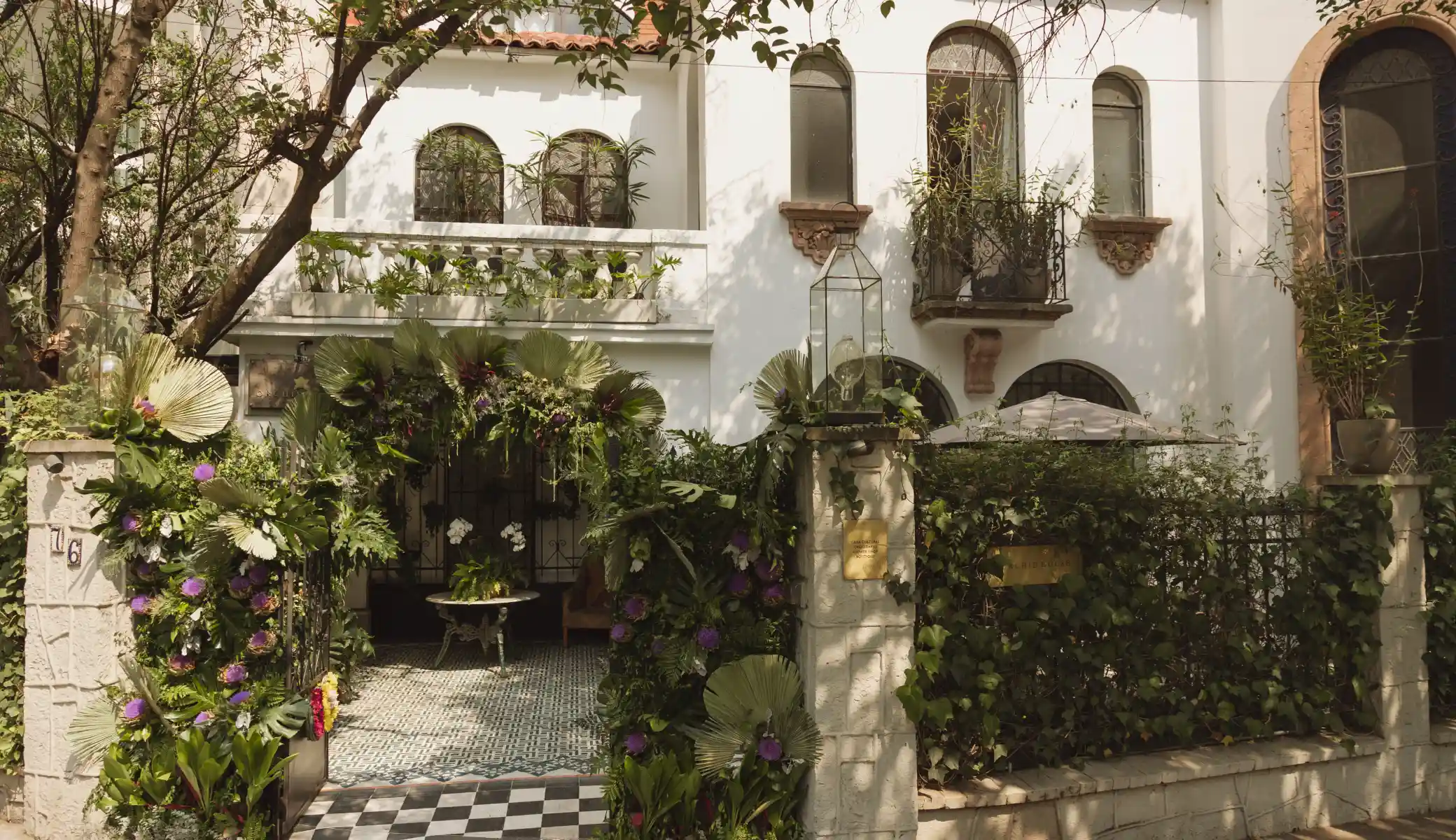 The entrance to Orchid House Polanco from the street: a white facade with arched windows, the doorway framed by an arch of flowers and palm leaves, and the chequered floor of the hallway
