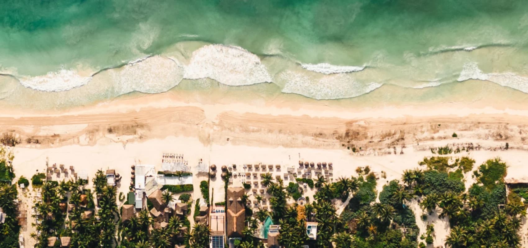 An aerial view of the Tulum coast: turquoise waves breaking on the sand, with palapas and palm trees below