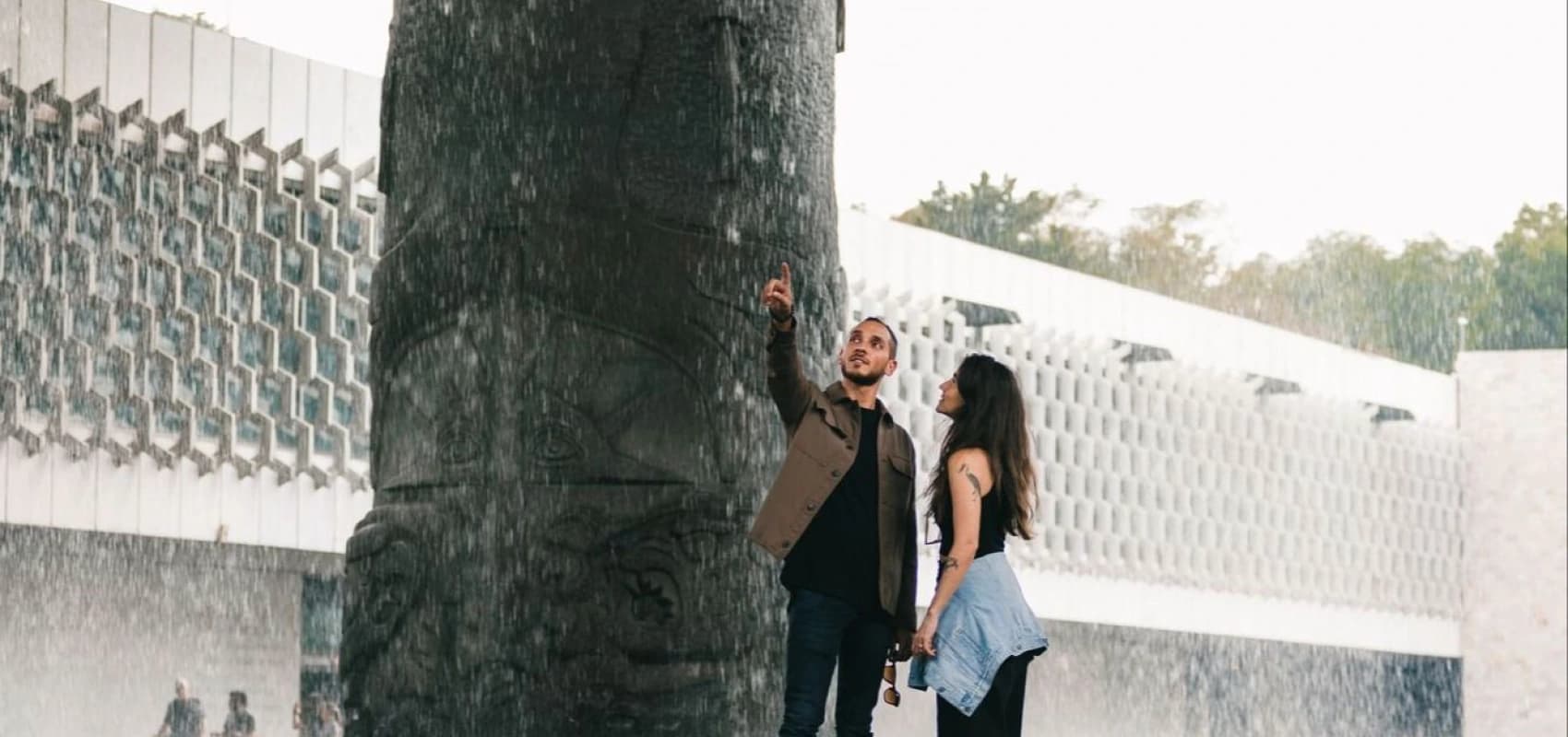A couple under the "El Paraguas" rain, reaching to touch the water falling from the carved column at the National Museum of Anthropology