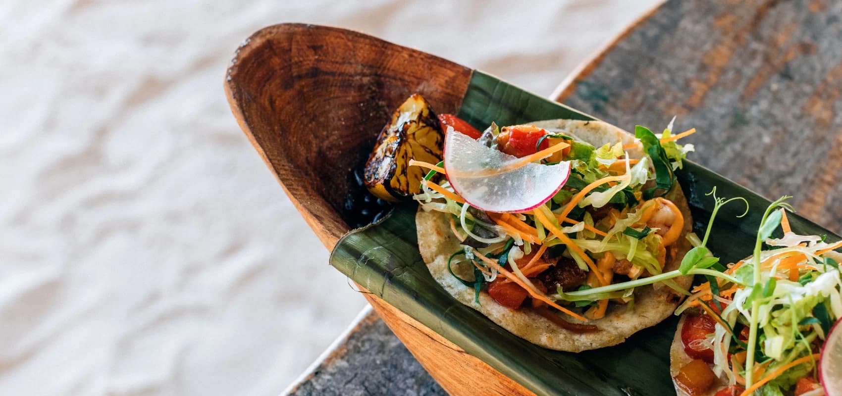 A seafood tostada on a banana leaf in a wooden dish, with grilled corn beside it and the beach sand in the background